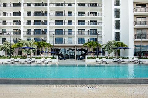 Apartments for Rent in Miami, FL - MB Station - Resort Style Pool With Lounge Chairs, Navy Umbrellas, And Modern Apartment Building With Balconies And Glass Doors.