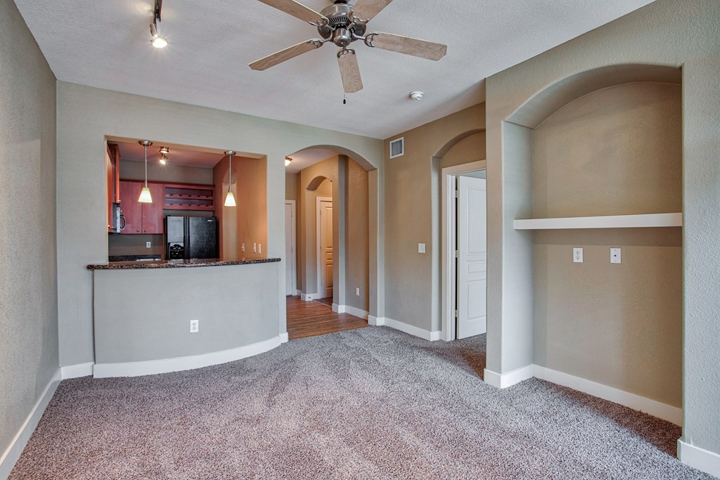 A living room with a ceiling fan and carpet and shelves  with kitchen in background