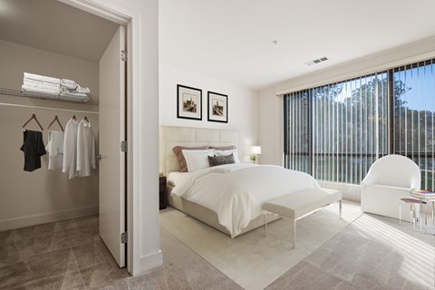 Novato, CA, Pet-Friendly Apartments - Millworks - Bedroom With White Bedding, Spacious Closet, Large Window With Vertical Blinds, White Accent Chair, Bench, And Carpeted Flooring.