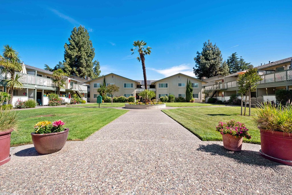 Apartments For Rent Gilroy, CA - Mission Park - Residential Courtyard In The Heart Of The Apartment Complex With Pebbled Paths, Potted Plants, Lush Green Grass, And Trees