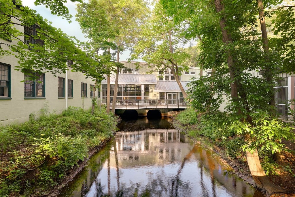 Scenic canal flowing under a building with a metal roof, surrounded by lush greenery.