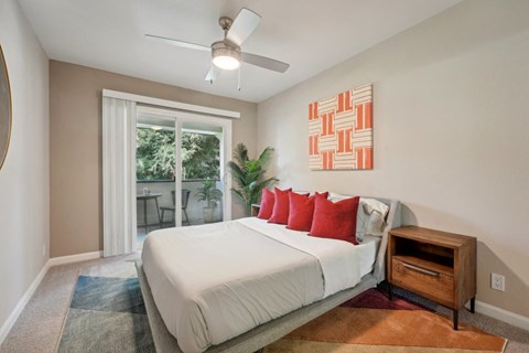 One-Bedroom Apartments in Fremont, CA - Northgate Savoy - Bedroom with Carpeted Flooring, Bed, Wooden Nightstand, Houseplant, Ceiling Fan, Beige Walls, Framed Mounted Mirror, and Sliding Glass Door to Balcony.
