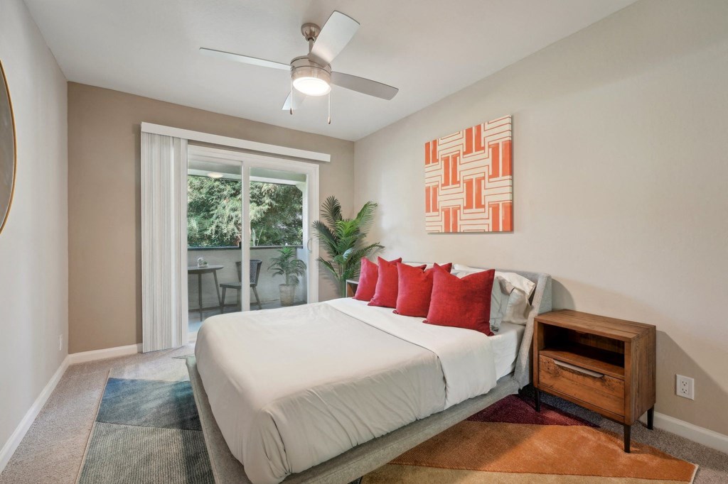 One-Bedroom Apartments in Fremont, CA - Northgate Savoy - Bedroom with Carpeted Flooring, Bed, Wooden Nightstand, Houseplant, Ceiling Fan, Beige Walls, Framed Mounted Mirror, and Sliding Glass Door to Balcony.