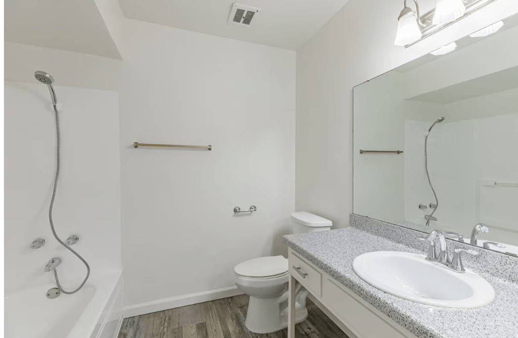 Three-Bedroom Apartments in El Cajon, CA - Ivanhoe Village - Bathroom with  Wood-Style Flooring, White Walls, Tub and Shower Combo, Granite-Patterned Countertops, and Large Mirror.