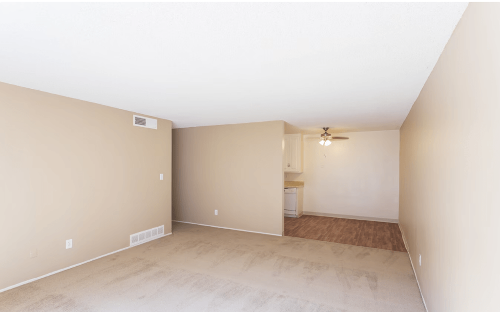 Two-Bedroom Apartments in El Cajon, CA - Ivanhoe Village - Unfurnished Living Room with Carpeted Flooring, Beige Walls, Ceiling Fan, and View of Kitchen.