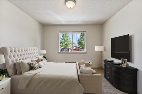 One-Bedroom Apartments in Vancouver, WA - Reflections at the Park - Spacious Bedroom With Tufted Headboard, Large Window, Neutral Carpet, And Sitting Area With Sofa.
