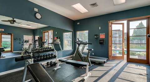 Coeur d'Alene, ID, Apartments - Rockwood Lodge - Fitness Center with Rubber Matting, Large Mirror, Dumbbells, Cardio Equipment, Ceiling Fan, Recessed Lighting, and a Doorway Leading to the Pool Area.