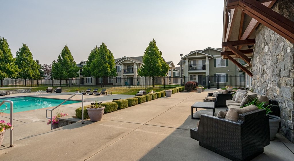 Apartments In Coeur d'Alene, ID, For Rent - Rockwood Lodge - Gated Resort-Style Pool With Expansive Sundeck, Lounge Seats, Manicured Hedgerow, Patio Area With Furniture, Trees, And Surrounding Apartment Buildings.