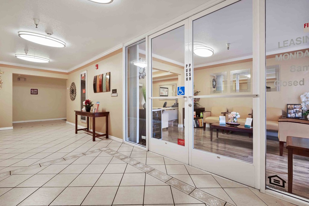 Two-Bedroom Apartments In Daly City, CA - Serra Commons - Leasing Office with Double Glass Doors, Wood-Inspired Flooring Inside, Waiting Area with Chairs, And Receptionist Desk.