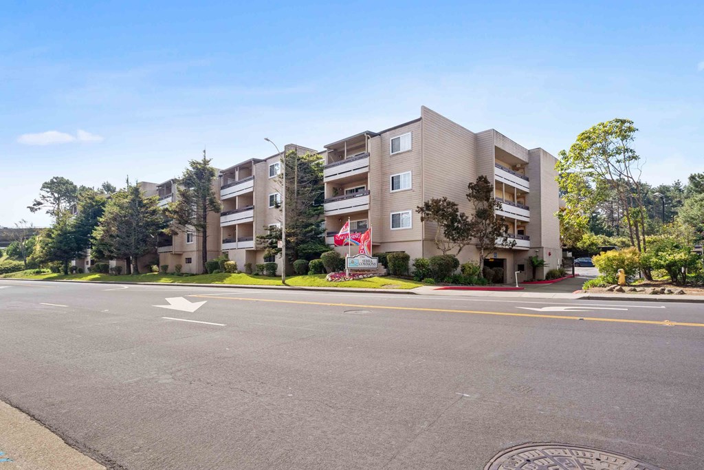 Apartments in Daly City, CA, for Rent - Serra Commons - Street View of Apartment Building with Property Signage, Flags, and Trees.