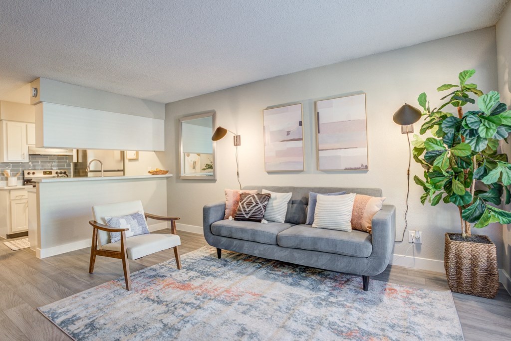 Studio Apartments in Glendale, AZ - Ardella on 67th - Living Room with Hardwood Floor, Area Rug, Couch, Armchair, Wall-Mounted Lamps, Wall Art, Houseplant, and View of Kitchen.
