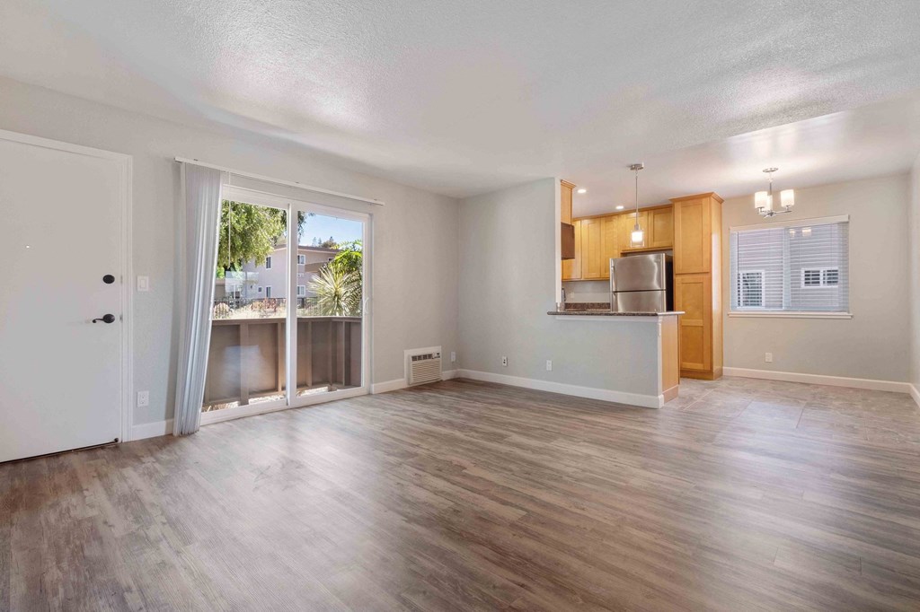 Dog-Friendly Apartments in Milpitas, CA - Spring Valley - Unfurnished Living Room Space with Wood-Style Flooring, Sliding Glass Door to Patio, White Walls, AC Unit, and View of Kitchen.