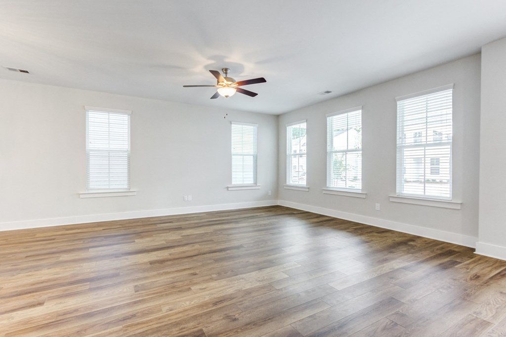Single Family Homes in North Charleston, SC - Sumner Village - Empty Living Room Area with Hardwood-Style Flooring, a Ceiling Fan with Lighting, and Multiple Large Windows with White Blinds.