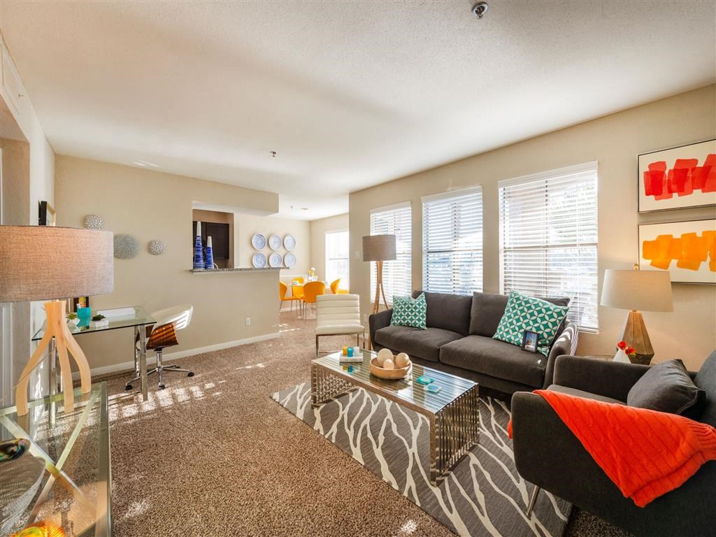League City, TX Apartments - Anchor at South Shore - Furnished Living Room with Carpet Flooring, Large Windows with Blinds, Stylish Decor, and a Dining Area Near the Kitchen