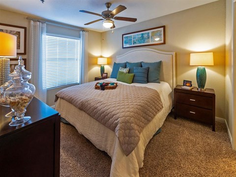2 BR Apartments in League City, TX - Anchor at South Shore - Bedroom with Carpet Flooring, a Bed, a Ceiling Fan, and a Large Window with Curtains and Blinds