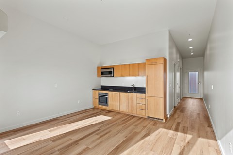 North East Portland, OR, Pet-Friendly Apartments - The Beverly - Open Apartment Kitchen And Living Area With Wood Style Flooring, Light Wood Cabinets, Black Countertops, And Bright Hallway.