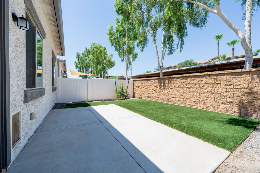 A concrete walkway leads to a house with a stone wall and green grass.