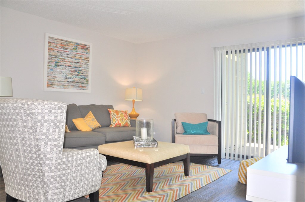 Bright living room with gray sofa, patterned armchair, beige ottoman coffee table, and colorful rug.