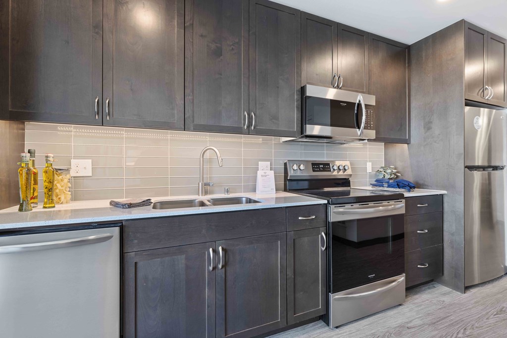 Apartments For Rent In Greenwood, WA - The Morrow - Fully Equipped Kitchen With Stainless Steel Appliances, Wooden Cabinets, Tile Backsplash, Granite Counters, And Vinyl Plank Flooring