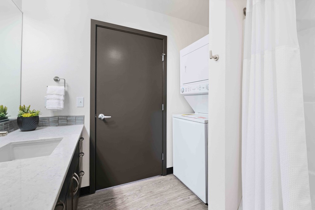 Greenwood, WA Apartments - The Morrow - Bathroom With In-Unit Washer And Dryer, Vinyl Plank Flooring, Granite Countertop, And Dark Brown Cabinets
