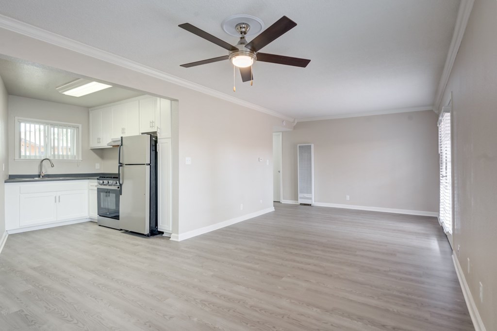 A spacious room with a ceiling fan and light fixture, a kitchen area with white cabinets and a stainless steel refrigerator, and a large window with blinds.