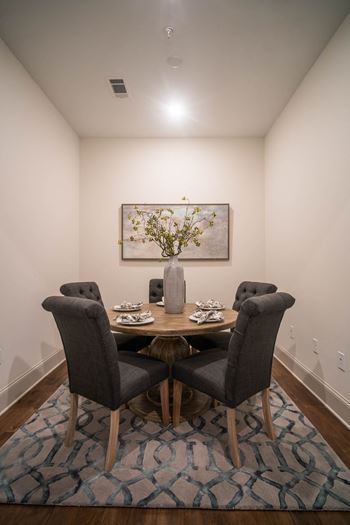 A compact dining area with a round table, four cushioned chairs, and a vase centerpiece on a patterned rug.