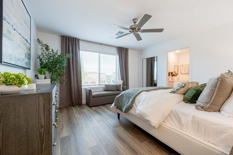 Townhomes in Tolleson, AZ, With In-Unit Washer and Dryer - Villas at 91st - Bedroom With Plush Bed, Ceiling Fan, Large Window With Curtains, Wood-Style Flooring, Dresser, Painting, and Bed Bench.