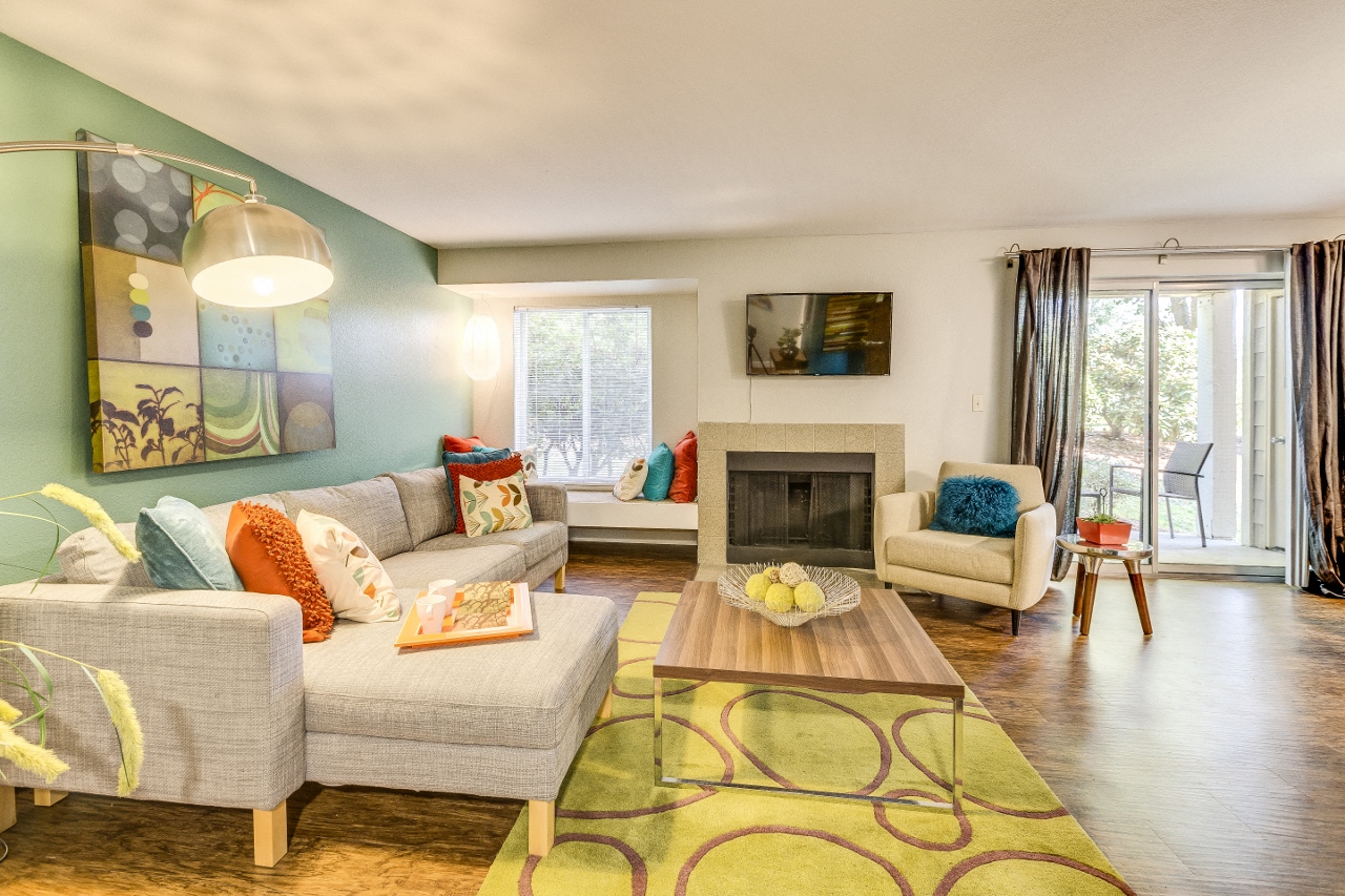 Apartments for Rent in Beaverton, OR - Spacious Living Room with Plank Flooring, Area Rug, Sectional Couch, Armchair, Wooden Coffeetable, Window-side Bench, Mounted TV, Fireplace, and Large Window with Curtains