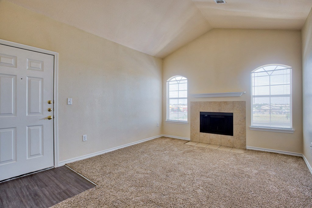 An empty living room with vaulted ceilings and fire pit and carpet and windows with blinds and wood floor at entrance of door