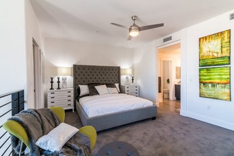 Summerlin, NV, Luxury Apartments Near Downtown Summerlin Mall - EVO Apartments - Bedroom With A Tufted Headboard, Nightstands, Table Lamps, Ceiling Fan, Accent Chair, And Connected Bathroom.