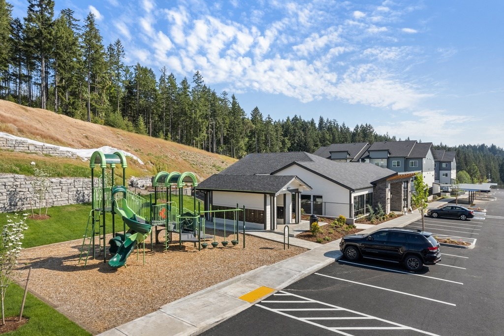 Belfair WA Apartments - Belfair View - a parking lot with a playground in the foreground and apartment buildings and trees In the background,