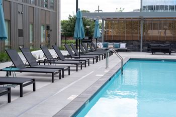 A pool with chairs and umbrellas is surrounded by a building.
