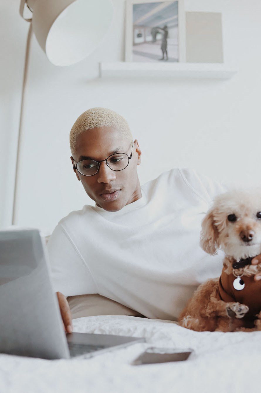 a man sitting on a bed with his dog while looking at a laptop
