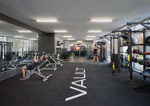 A gym with a man working out and a woman walking in the background.