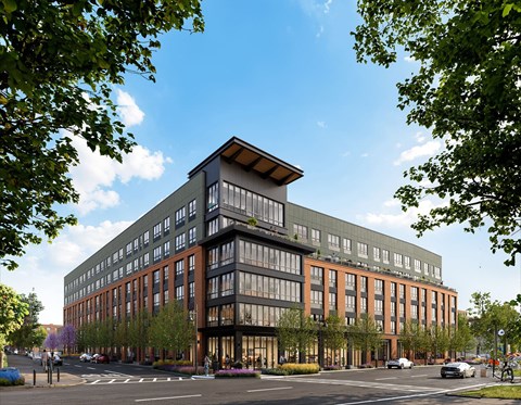 A modern multi-story building with a mix of glass and brick exterior is surrounded by trees and has a street in front of it.