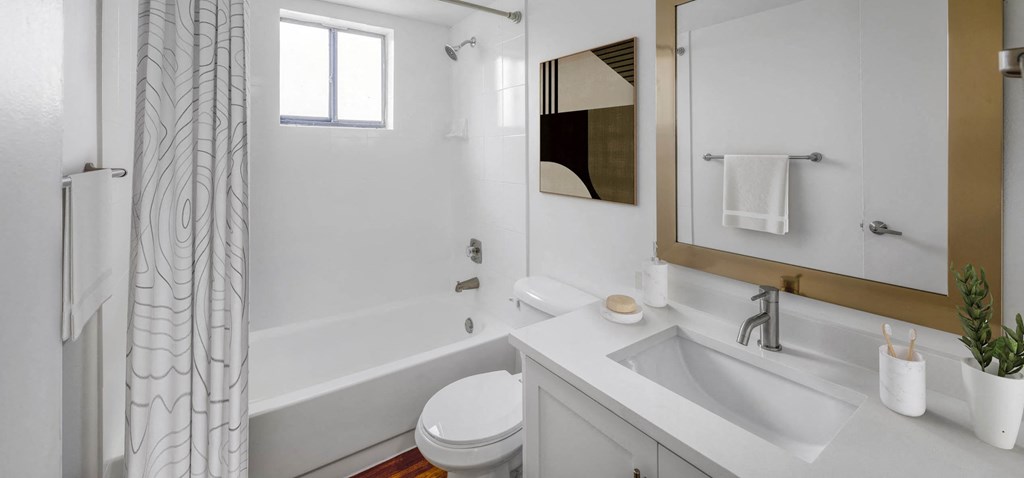 Bright, modern all-white bathroom with tub, toilet, vanity, and gold-framed mirror.