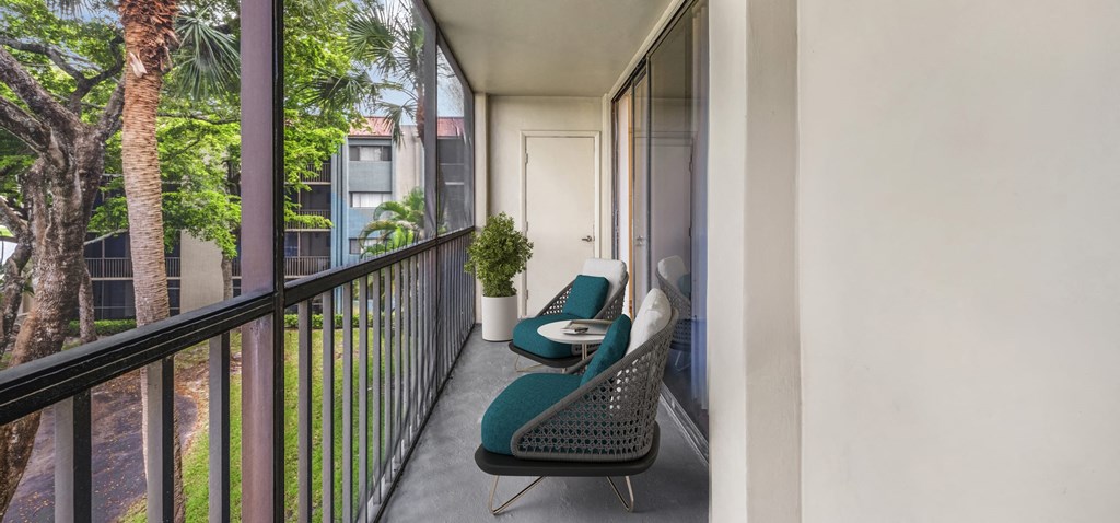 Apartment balcony with teal chairs, small table, potted plant, and view of trees.