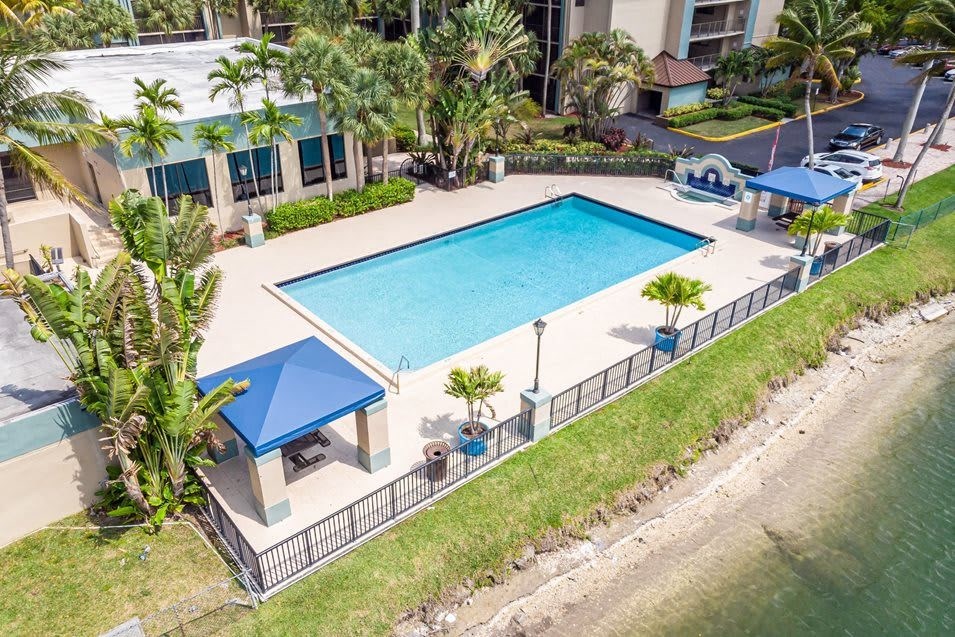 Aerial view of a rectangular swimming pool with blue water, surrounded by palm trees, a blue-roofed gazebo, and a lake.
