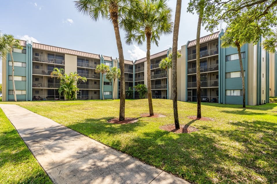 Exterior of a multi-story apartment building with balconies, surrounded by palm trees and green lawn.