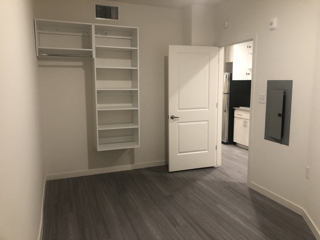 Dog-Friendly Apartments in Downtown Phoenix, AZ - VB on 12th Street - an Empty Unfurnished Room with Hardwood-Style Flooring, Wall-Mounted White Shelves, an Electrical Box, and a View Into the Kitchen.