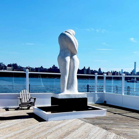 A sculpture of a nude woman stands on a dock with a chair and a cityscape in the background.