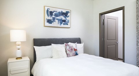 A bedroom with a white bed, a white nightstand with a lamp, and a framed blue and white painting above the bed.