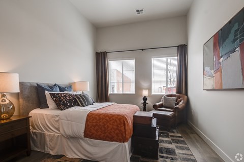 One Bedroom Luxury Apartments in Greeley CO - The Shelby - Furnished Bedroom with a Large Bed, a Leather Chair, Two Windows, White Walls, a Wooden Nightstand, and Wood-Style Flooring
