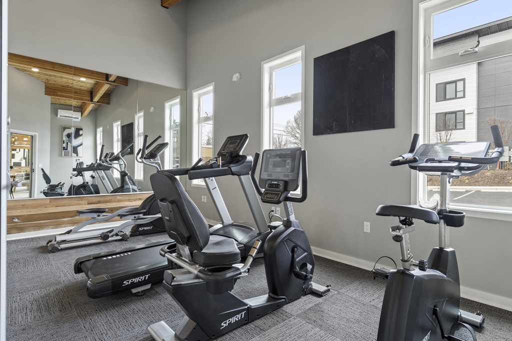 Tacoma, WA Apartments - Pacific Ridge - Fitness Center with Carpetted Floor, Cardio Machines, Large Windows, TV, High Ceilings, and Large Mirrors