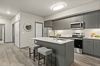 Apartments In Port Orchard For Rent - Belfair View - A Kitchen With Gray Cabinets, White Countertops, Stainless Steel Appliances, And Wood Flooring.