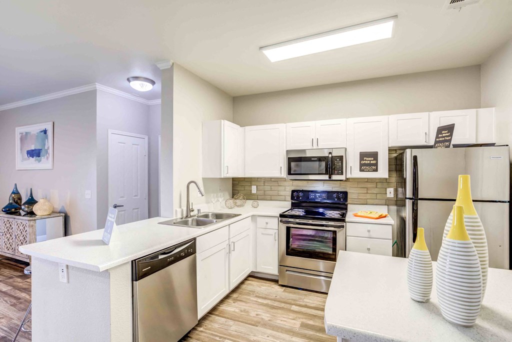 Two-BR Apartments In Henderson, NV - Avalon At Seven Hills - Modern Kitchen With White Cabinetry, White Counters, Stainless Steel Appliances, Tile Backsplash, And Wood Plank Flooring