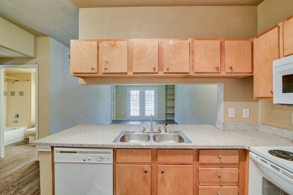 A living room and kitchen with wood flooring and wood cabinets with white dishwasher and white microwave and stove top with oven and steel sink