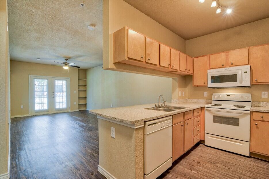 A living room and kitchen with wood flooring and wood cabinets with white dishwasher and white microwave and stove top with oven and steel sink