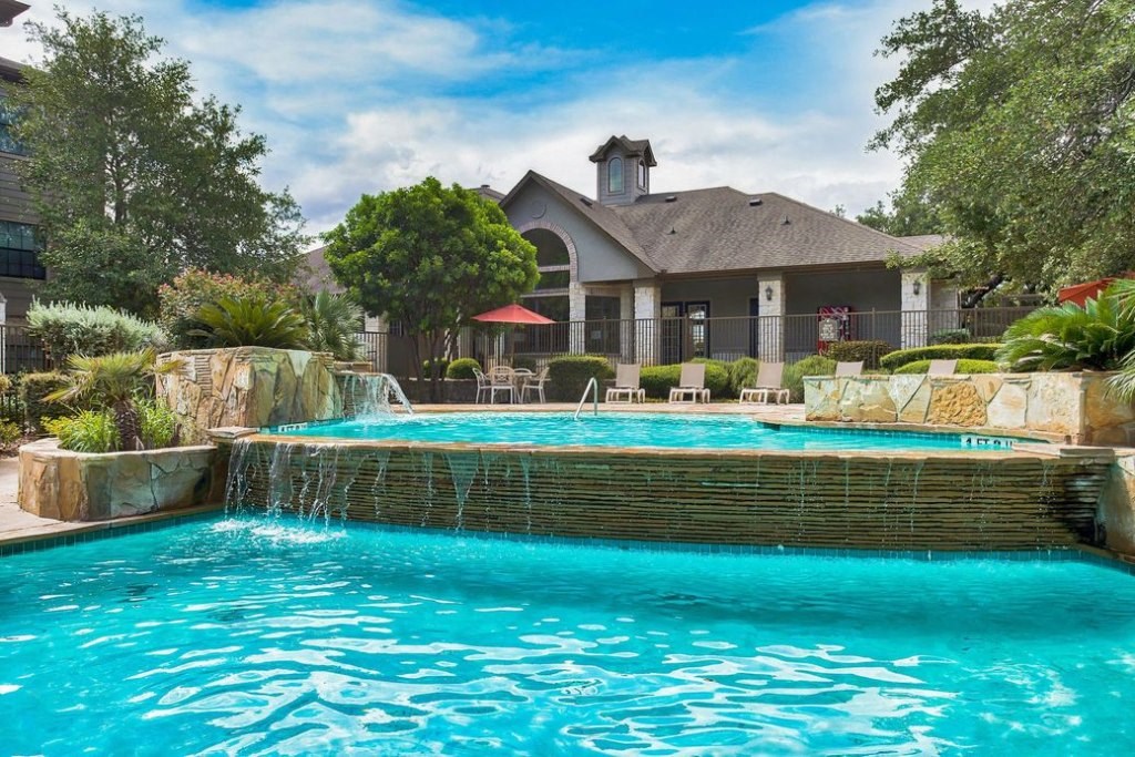 A pool with a water fall surrounded by rock floors and beach chairs and apartments
