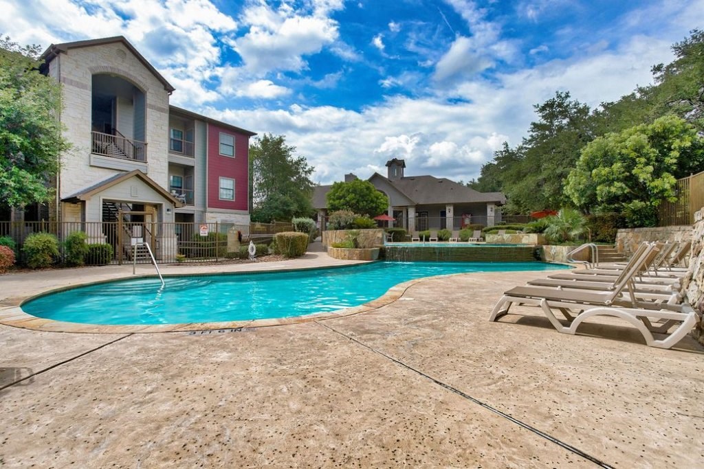 A pool with a water fall surrounded by rock floors and beach chairs and apartments